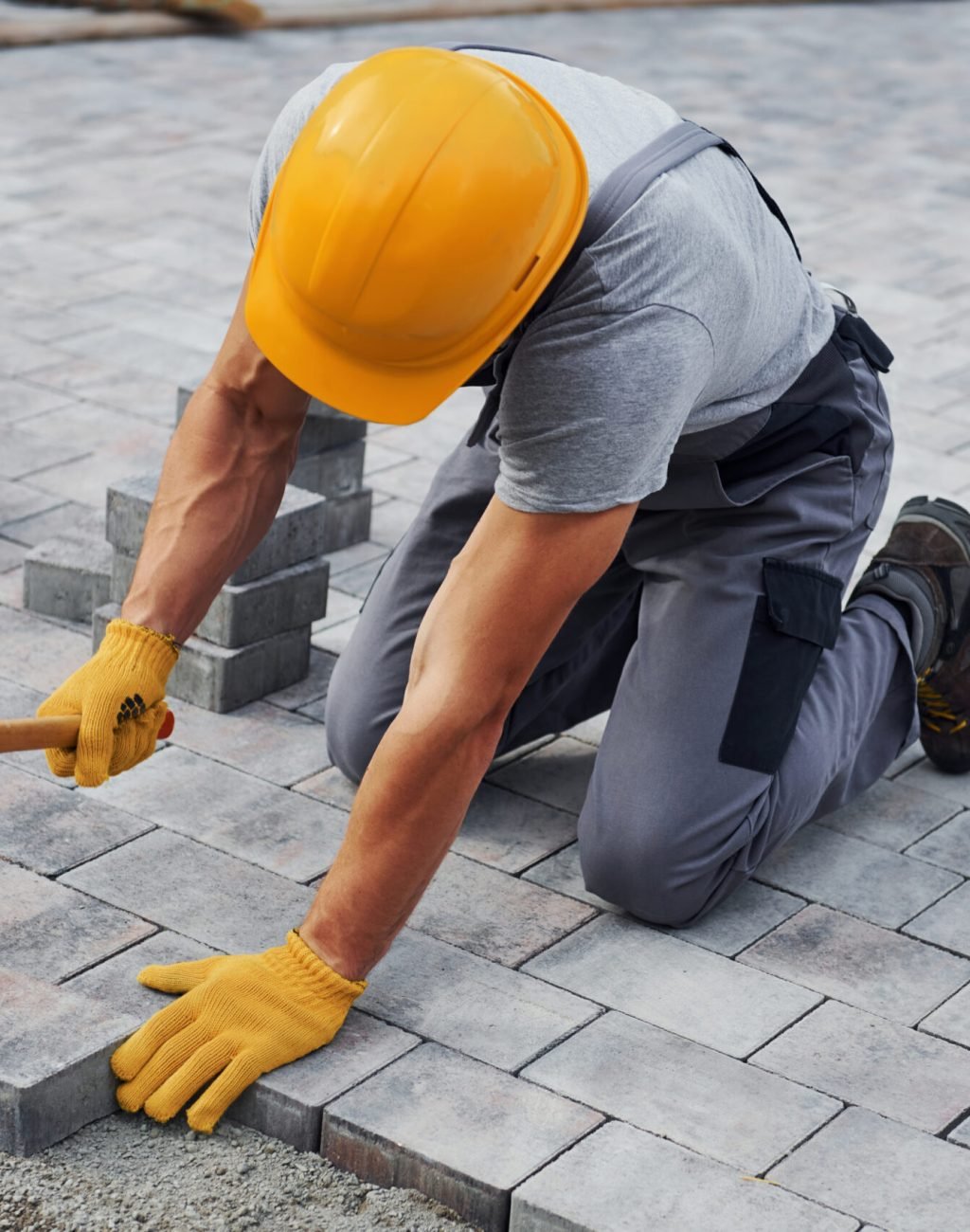 Uses a rubber hammer. Male worker in yellow colored uniform have job with pavement.